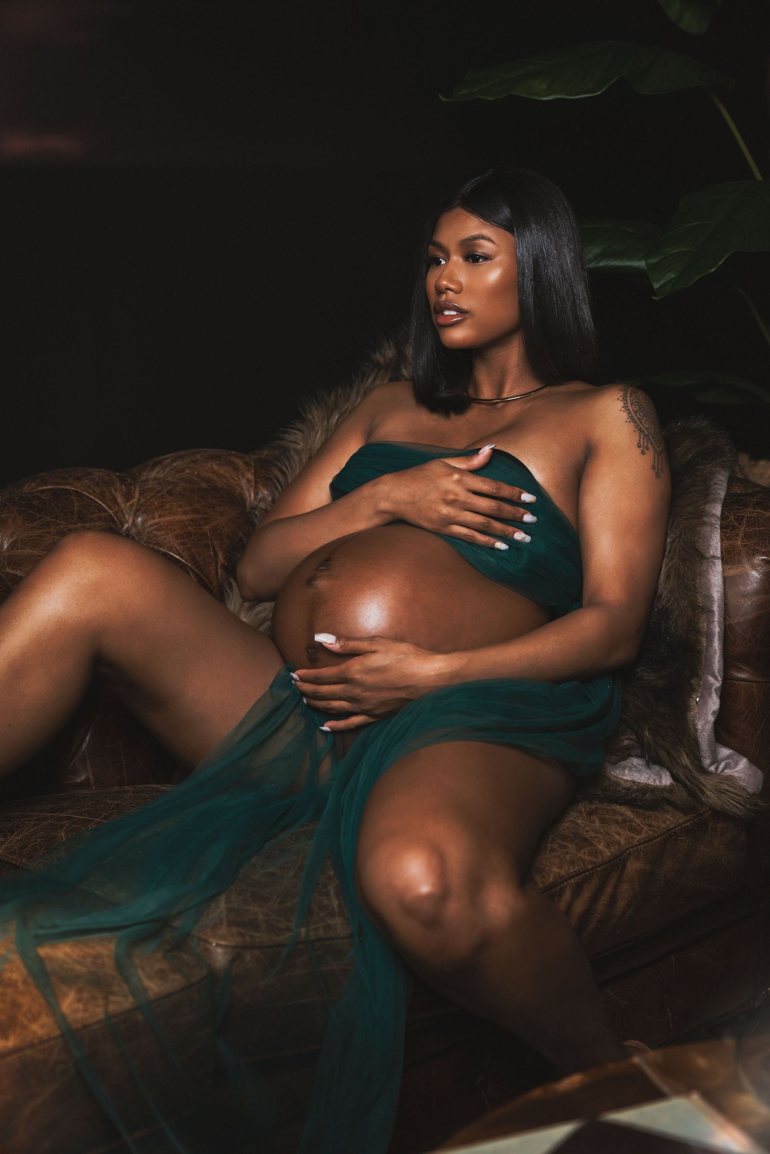 Pregnant Black woman in emerald green fabric reclining on vintage sofa in dramatic lighting for her maternity session in Cincinnati