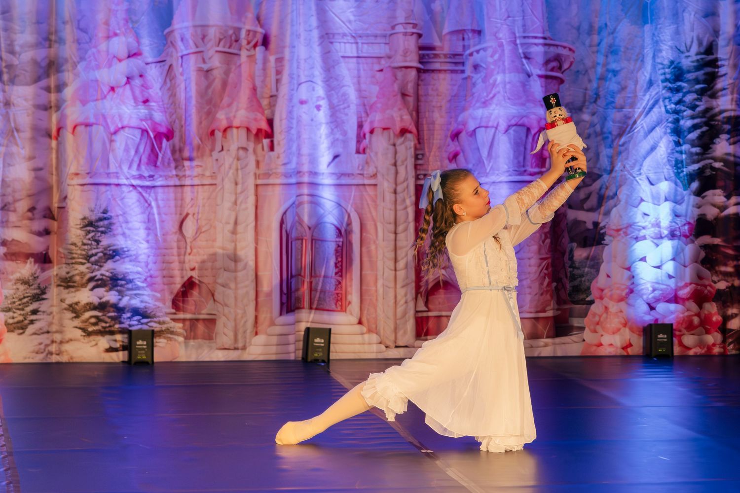 Ballet dancers perform gracefully on stage with elegant costumes against a backdrop illuminated in purple and blue lighting.