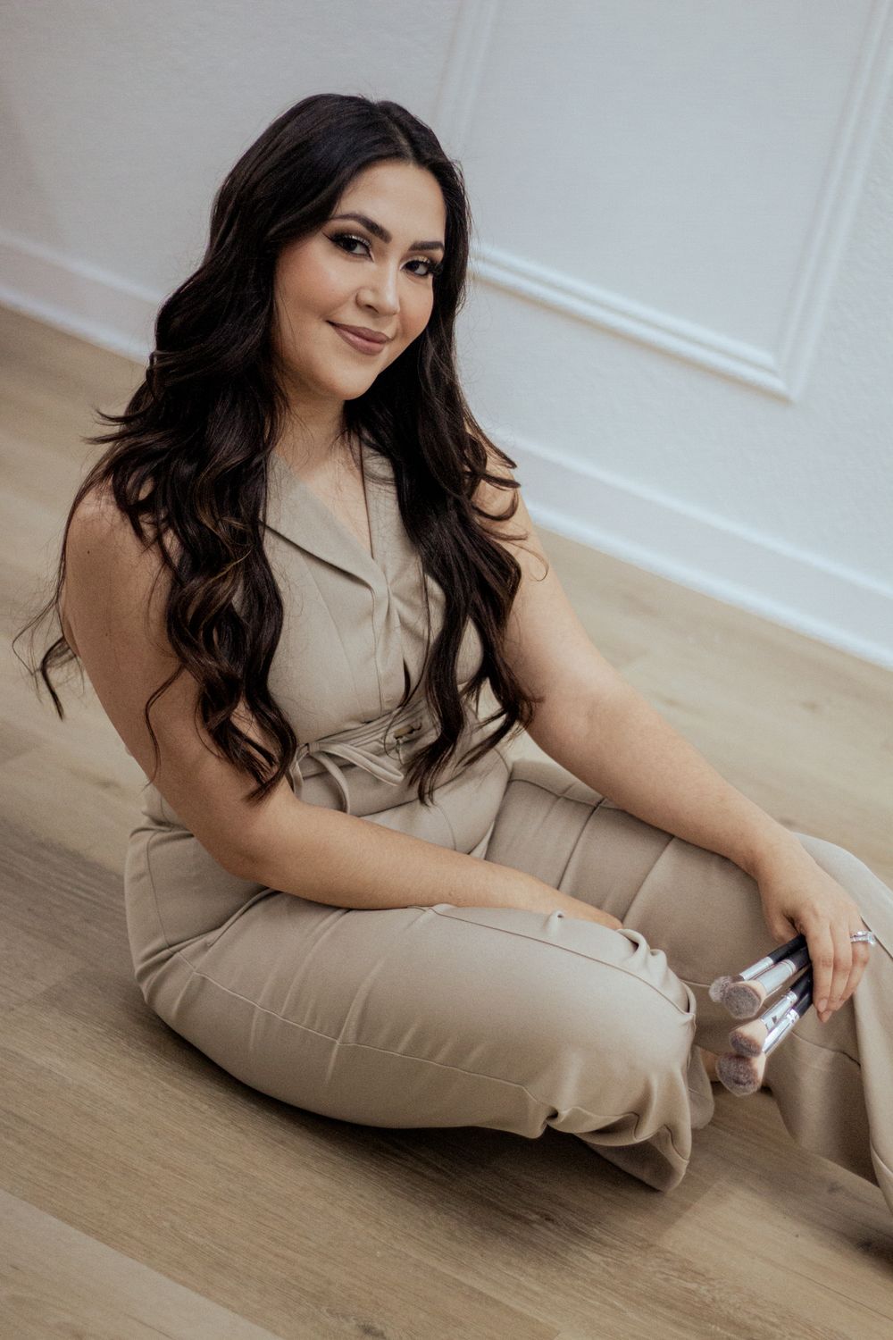 A woman in a neutral-toned jumpsuit sits casually on a hardwood floor beside white walls.