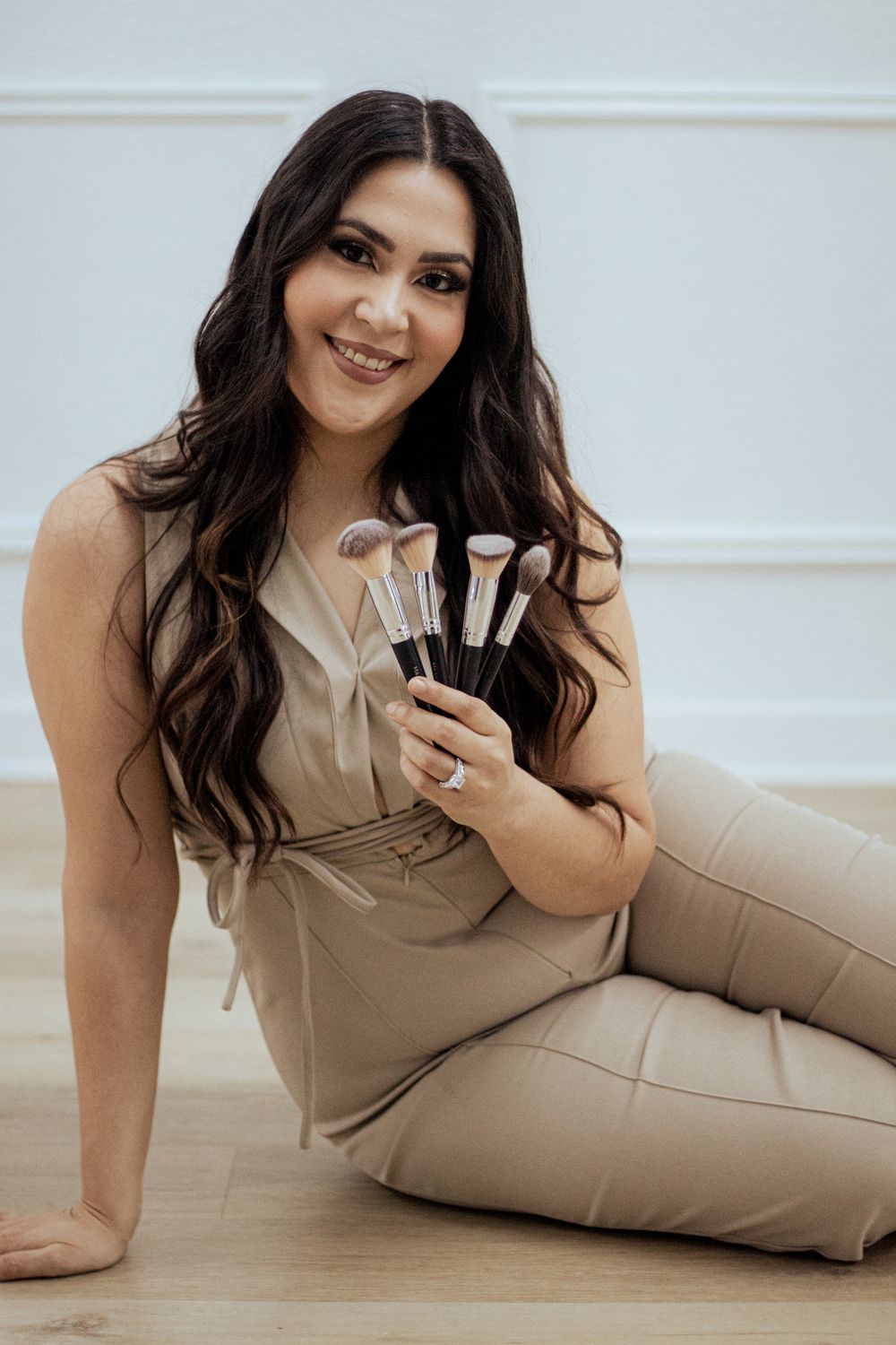 A beauty professional in beige clothing sits on hardwood floor holding makeup brushes with a bright smile.