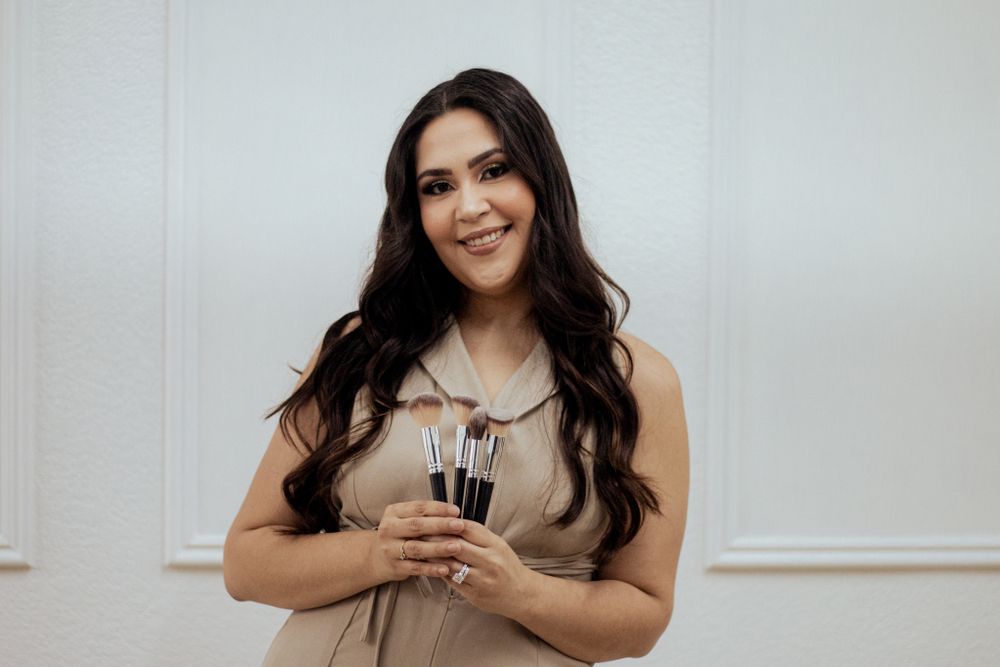 A person with long dark hair wearing a beige dress holds makeup brushes while standing against a white wall.