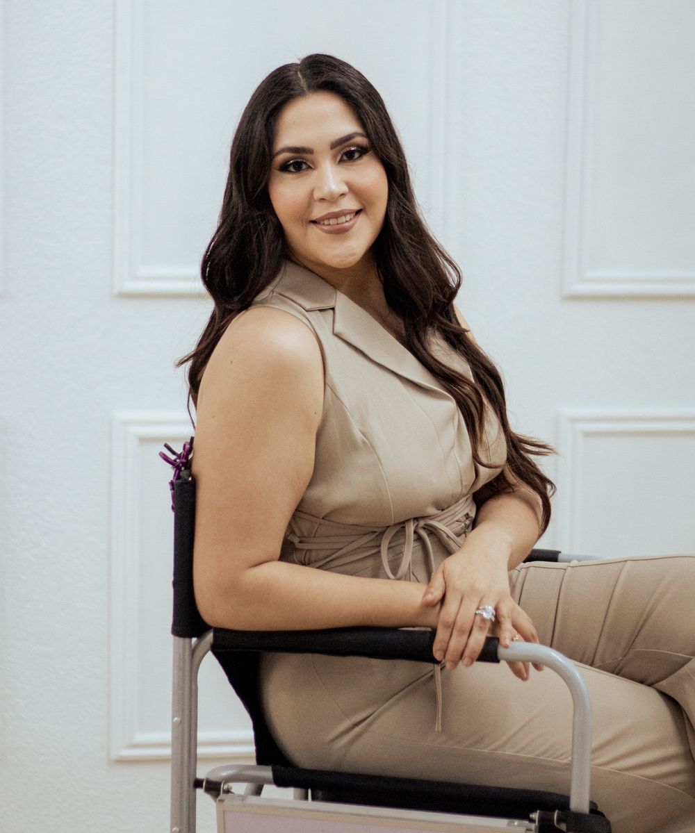 Professional portrait of someone seated in a beige sleeveless outfit against a white wall.