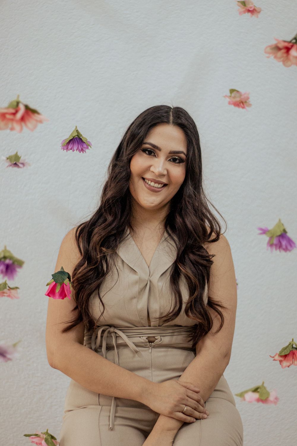 Portrait against floral backdrop wearing beige jumpsuit with wavy dark hair and delicate floral decorations.