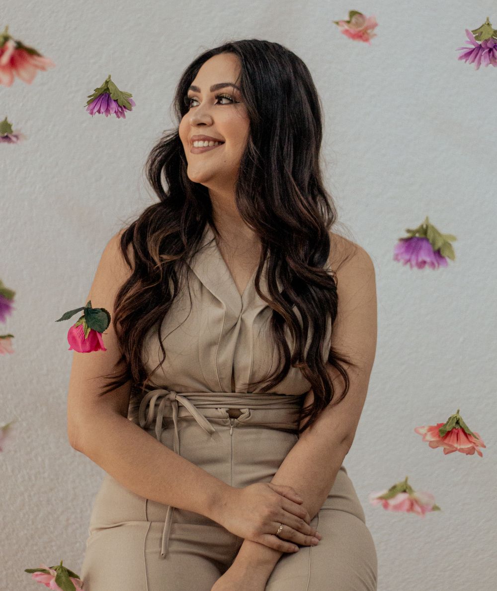 Smiling figure in a beige dress poses against white wall decorated with scattered pink and purple flowers.