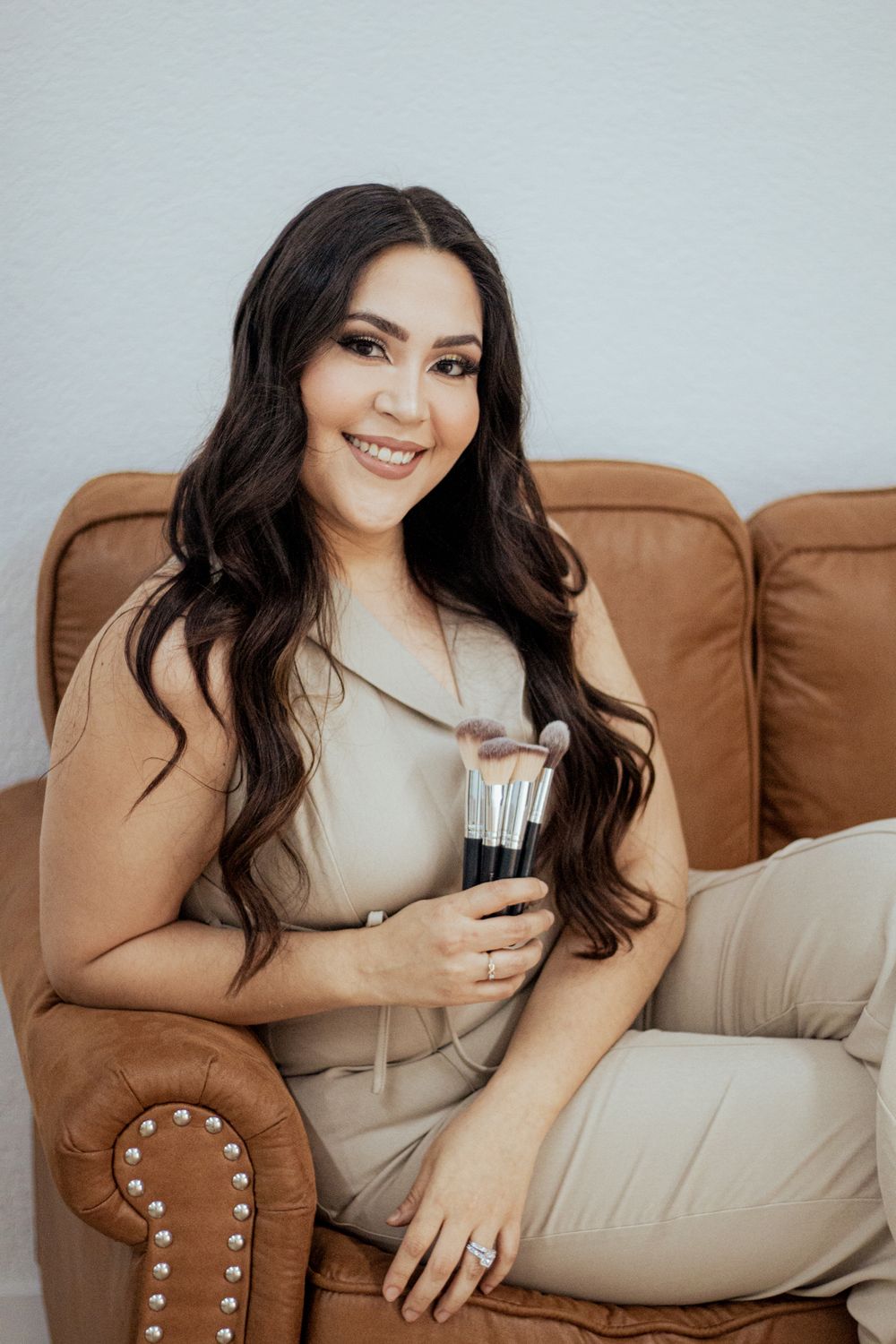 Woman in neutral beige outfit relaxes on brown leather armchair while holding makeup brush.