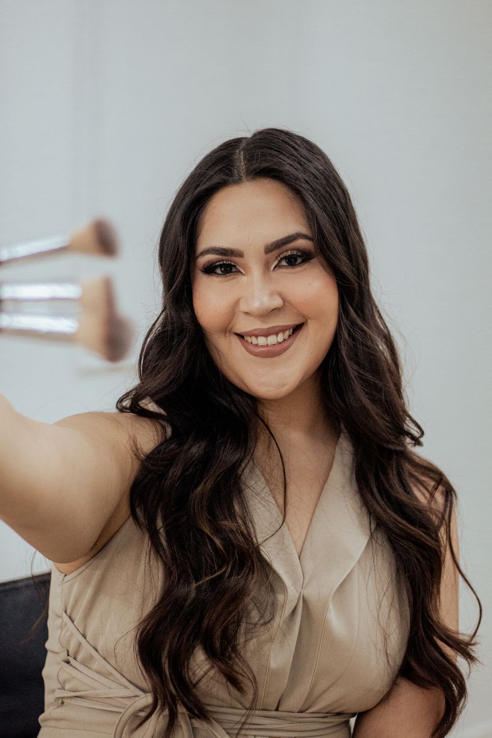 A brunette woman in a beige wrap top smiles warmly while holding a makeup brush against a light background.