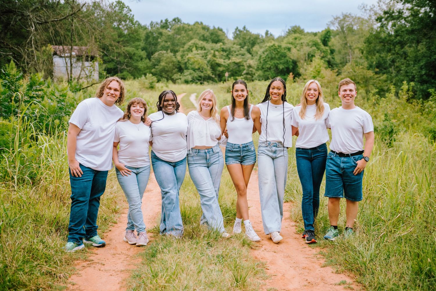 A group of friends in white shirts and jeans pose together in a natural outdoor setting on a dirt path.