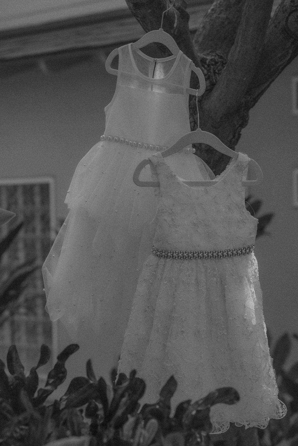 Two white lace flower girl dresses with tulle skirts and embellished waistbands hang on display in black and white.
