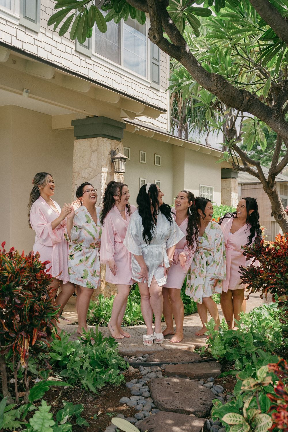 A group wearing matching floral robes poses together in a lush tropical garden setting with white brick walls.