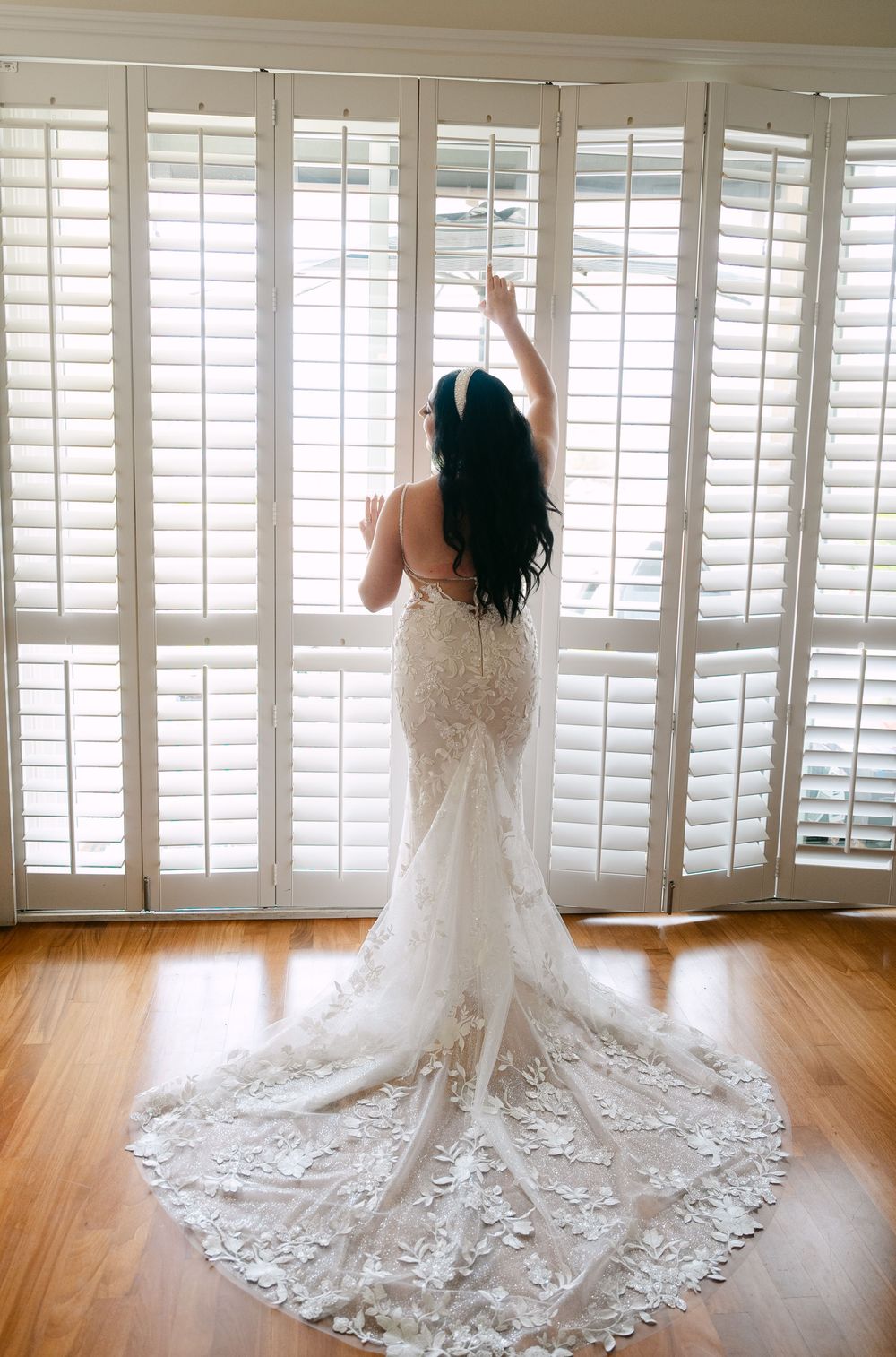 A stunning bridal gown with intricate lace details and long train is showcased in a sun-lit room with white shutters.