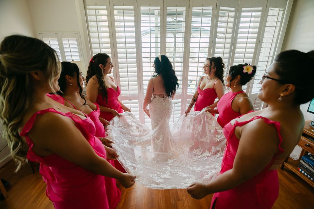 A group of people in pink dresses gather near white shuttered windows in a bright indoor setting.