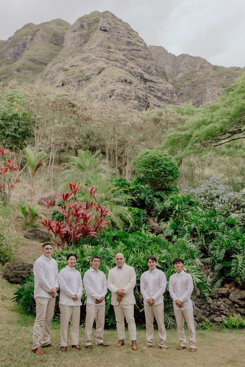 Wedding party in light beige suits posing against dramatic mountain backdrop in tropical garden setting.