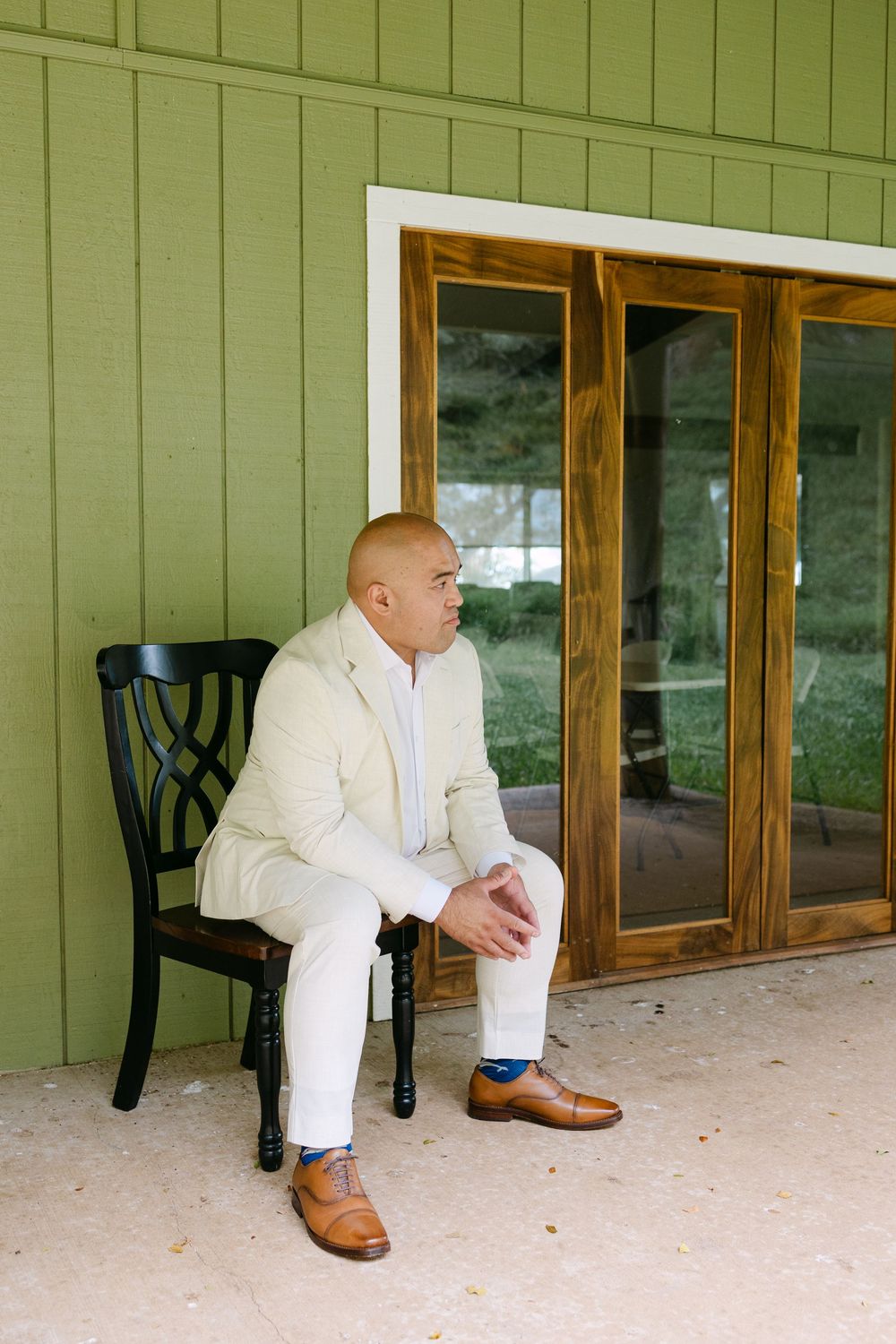 A person in a white suit sits thoughtfully on a wooden chair on a porch with green walls and wooden windows.