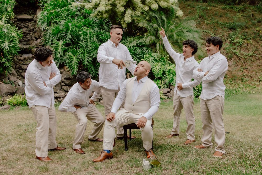 Groomsmen in light beige suits celebrate joyfully on a green lawn during an outdoor wedding photoshoot.