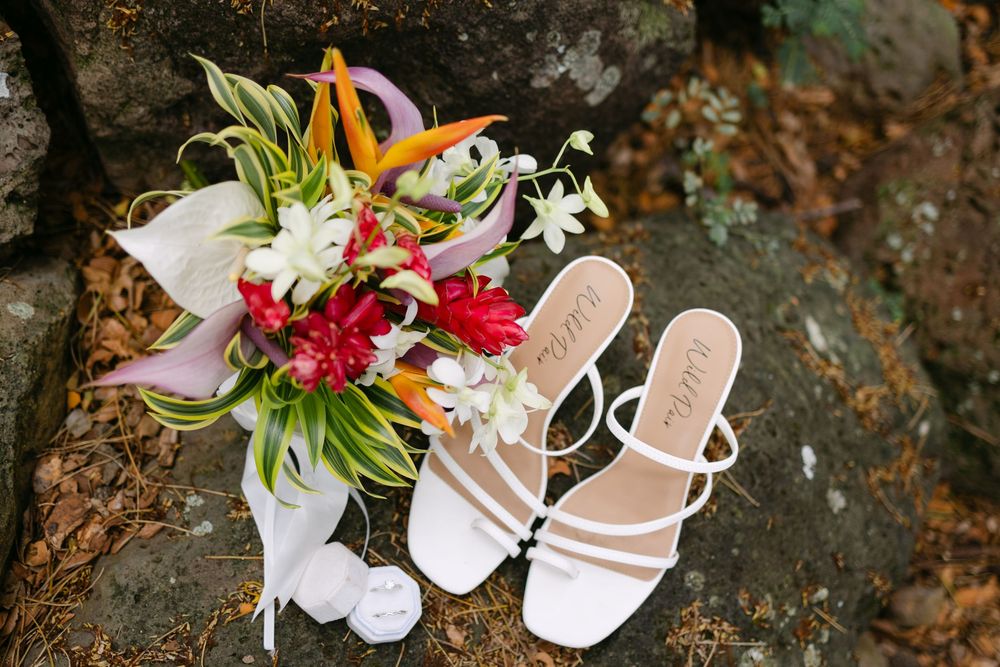 Elegant wedding details including bridal sandals, floral bouquets, and delicate accessories arranged on rustic textured surface.