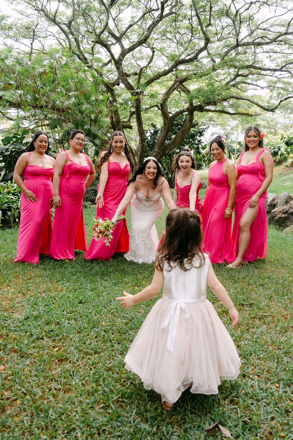 Bridesmaids in bright pink dresses gather around bride in white gown on grassy lawn under tropical trees.