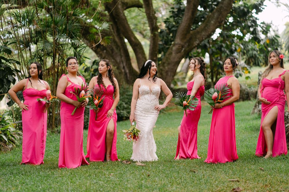 A bridal party in bright pink bridesmaid dresses poses together on a grassy lawn during an outdoor wedding celebration.