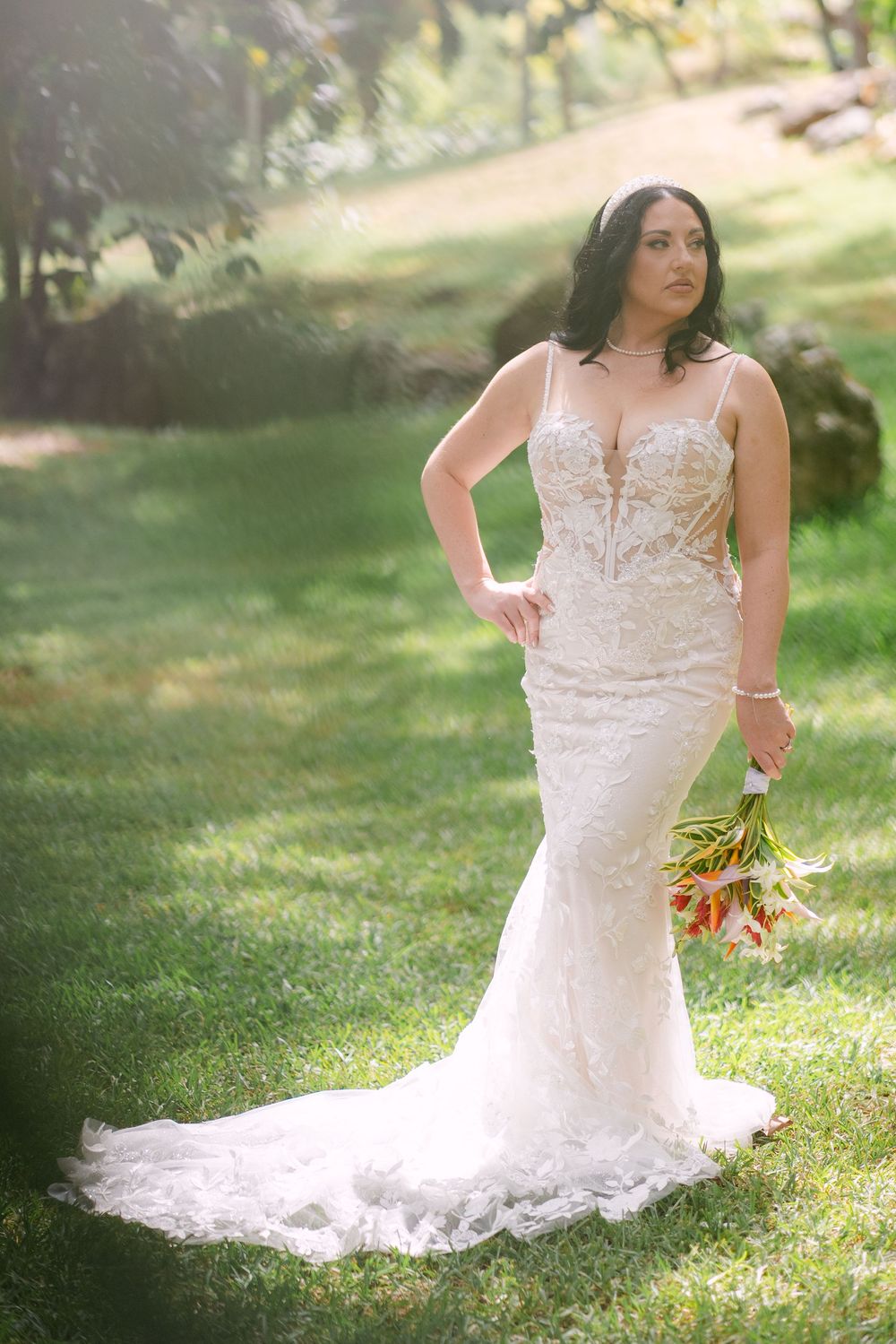 A bride in a fitted lace wedding gown poses elegantly in a sunlit garden while holding a floral bouquet.