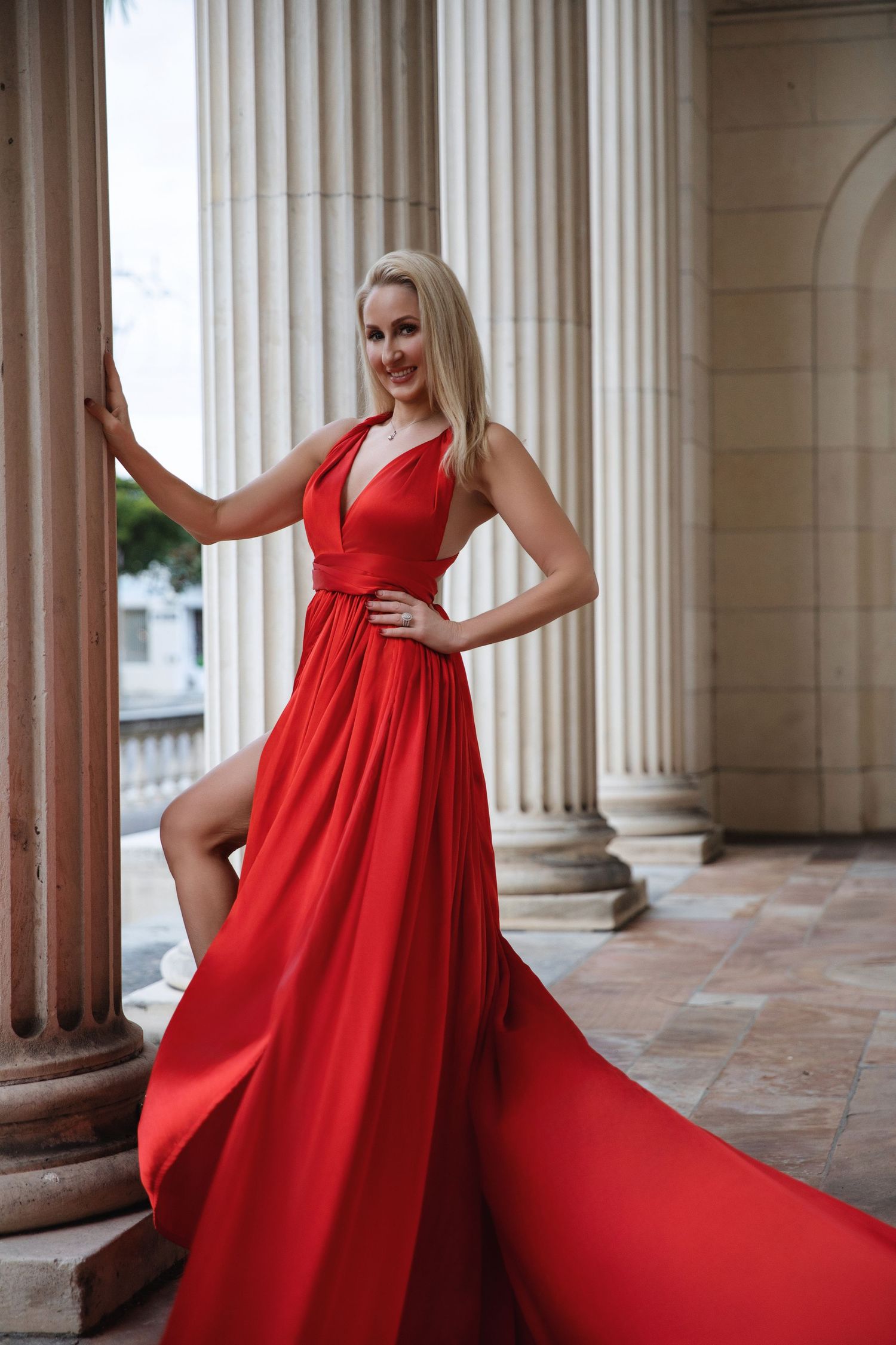 A flowing red gown billows dramatically between classical stone columns in an elegant architectural setting.