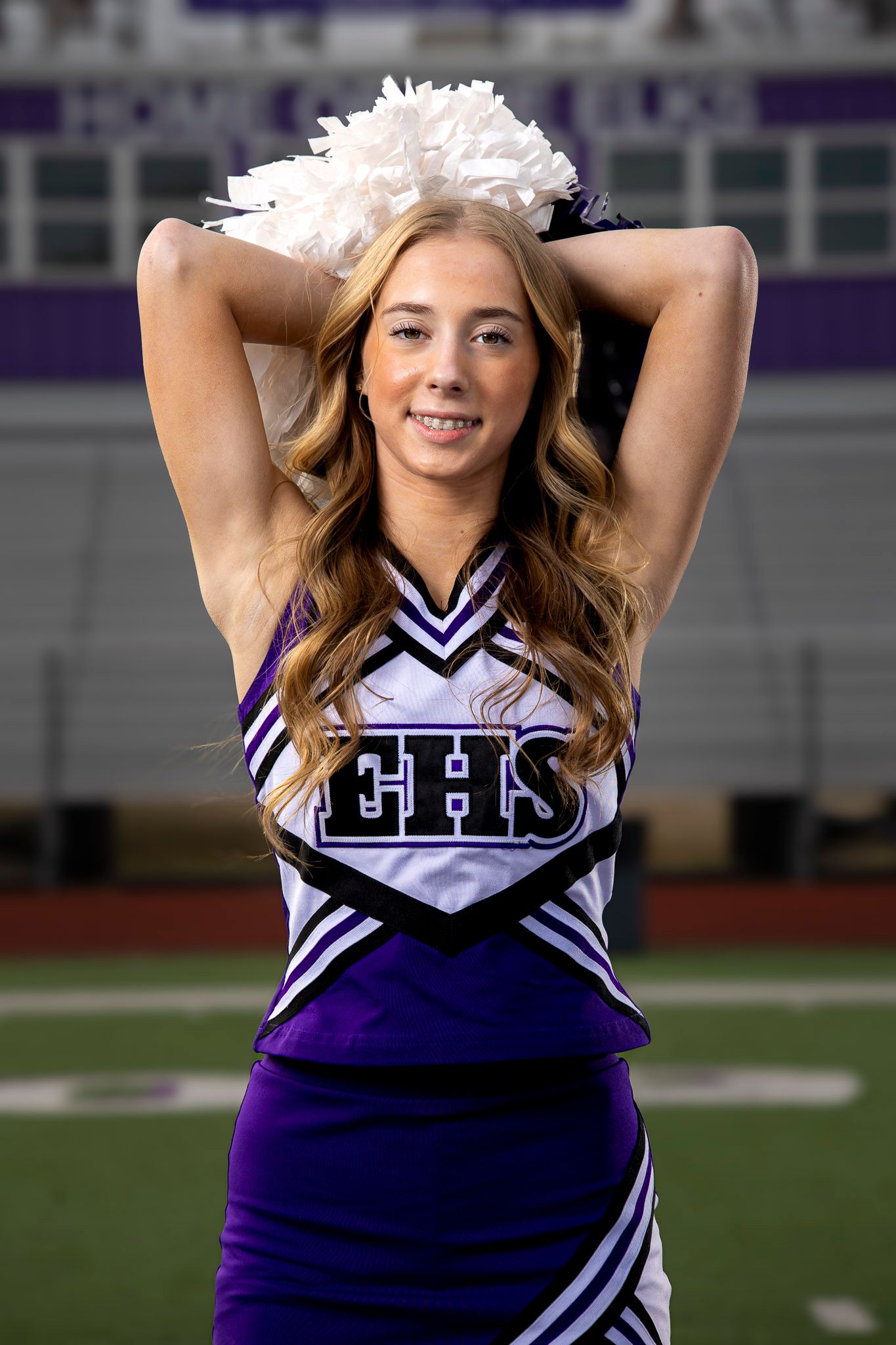 Cheerleader in purple and white uniform with pom poms poses on football field at high school stadium.