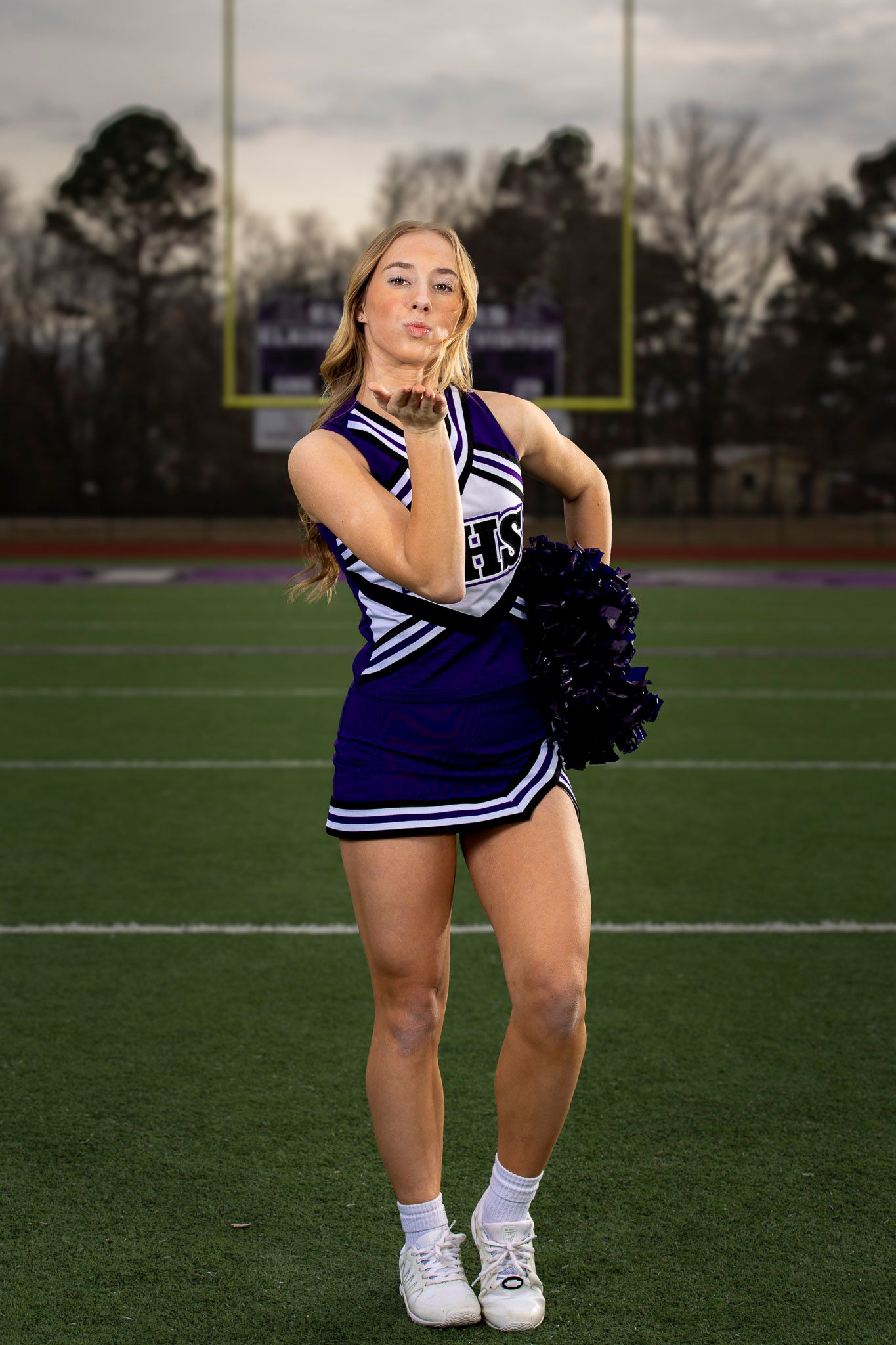 A cheerleader in a blue and white uniform performs a dynamic pose on a sports field at dusk.