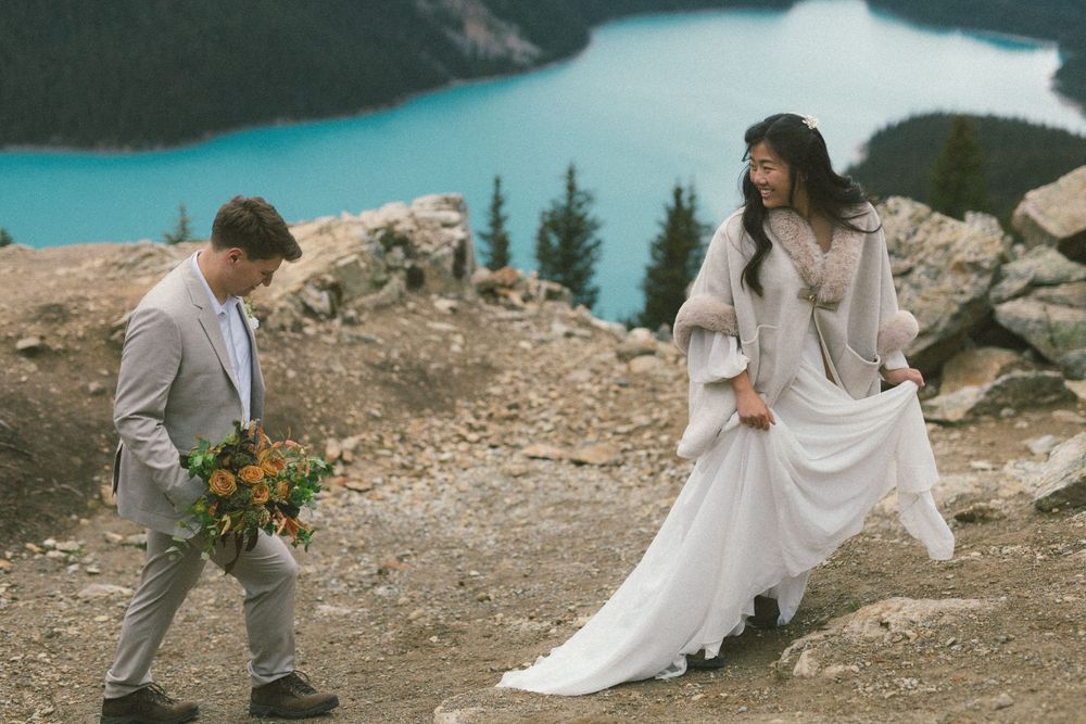 bride laughs and she and groom walk along rocky outcrop above a mountain lake in Banff