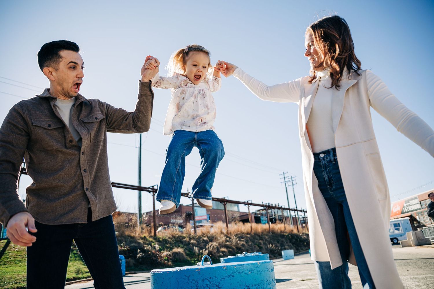 A child jumps with joy while being supported by two adults on a bright sunny day outdoors at Camp North End