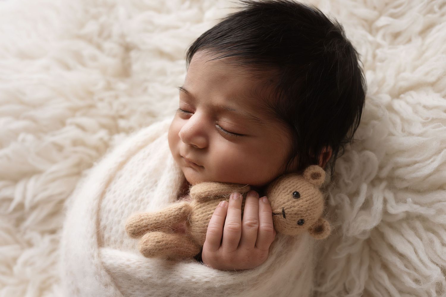 A sleeping newborn baby rests peacefully on soft white fur with a tiny teddy bear prop.
