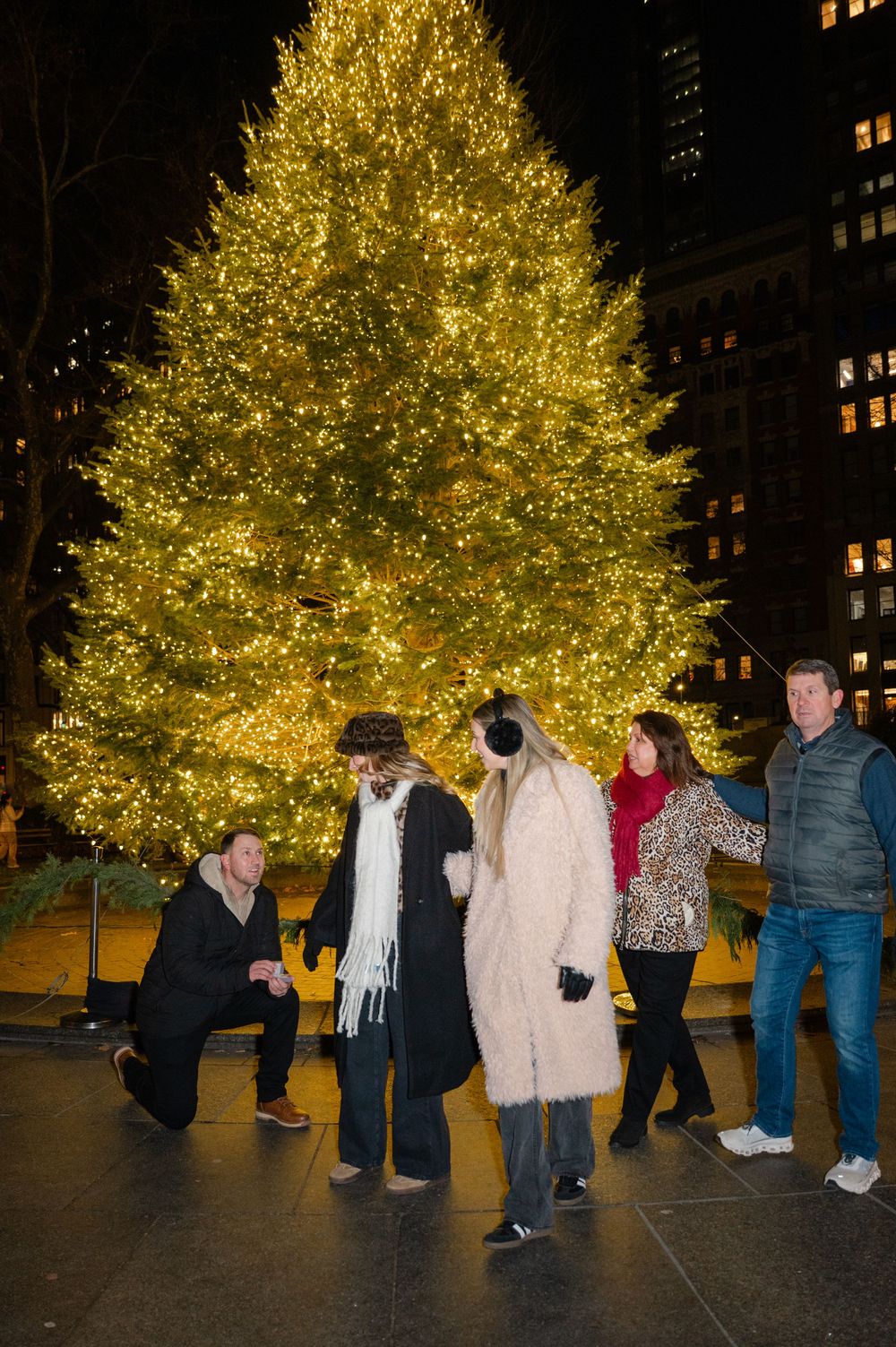 A man kneels down to propose to his girlfriend after her family takes a photo together in front of a lit up Christmas tree in NYC.