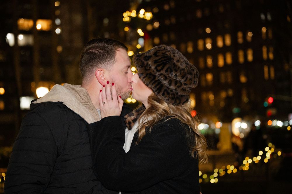A romantic couple shares a tender kiss at night with glowing city lights in the background.