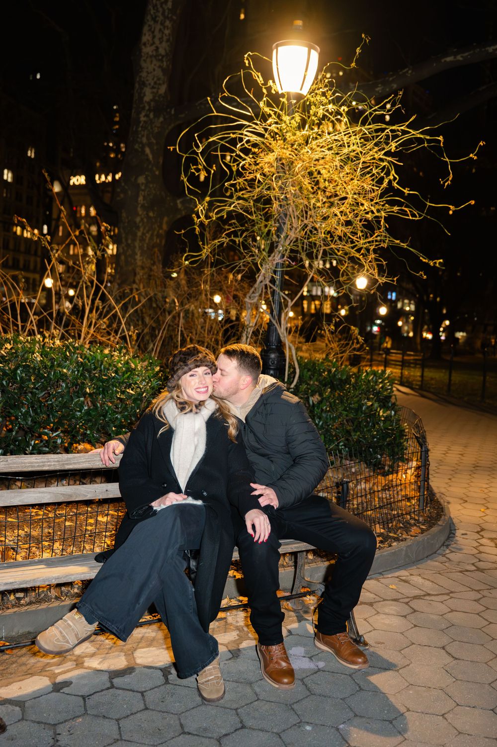 A couple sits together on a park bench at night with decorative lights glowing in the background.