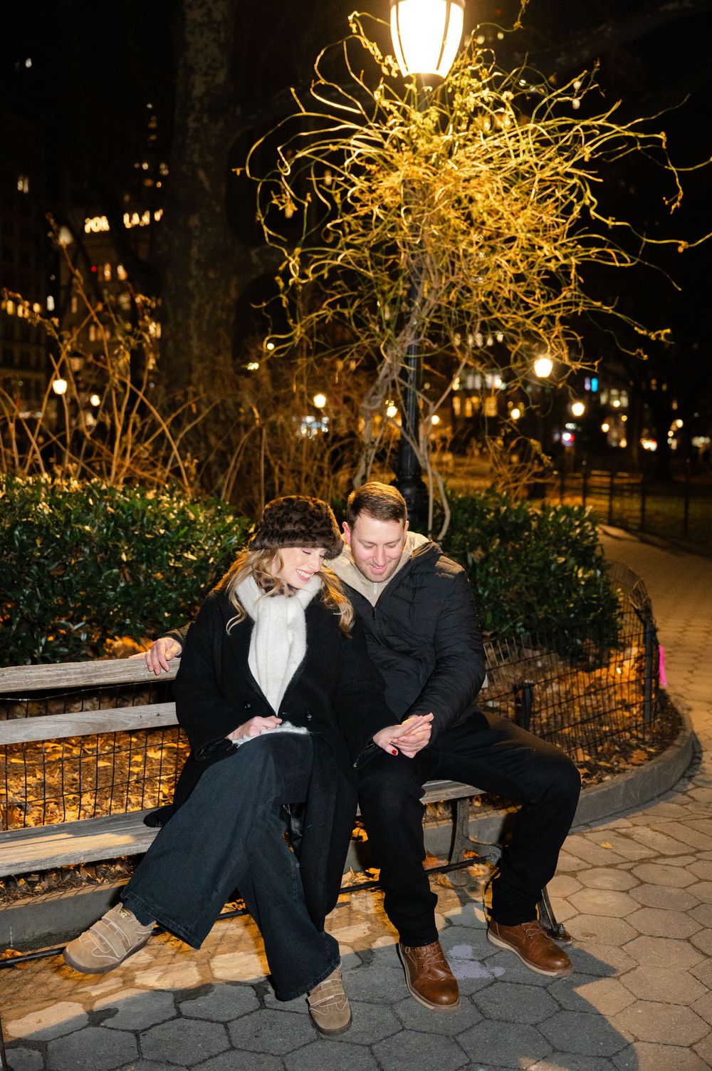 A newly engaged couple admires her engagement ring in the moonlight on a park bench in NYC.