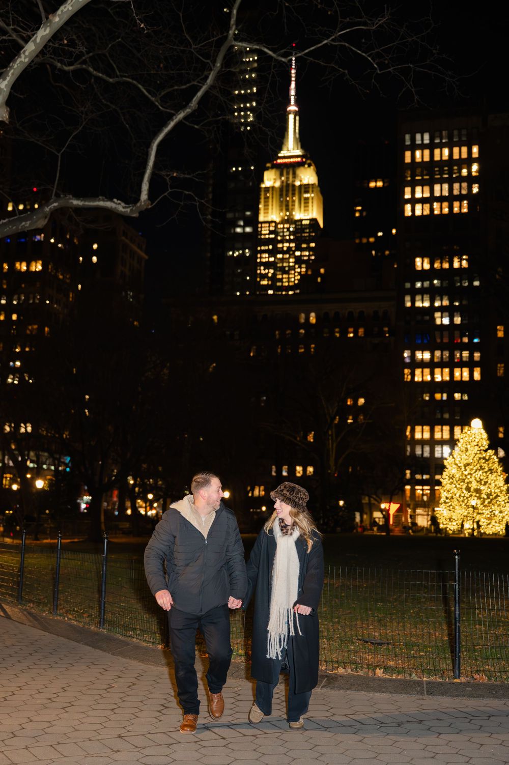 Two people walking along an illuminated path with city skyscrapers and twinkling lights in the background.