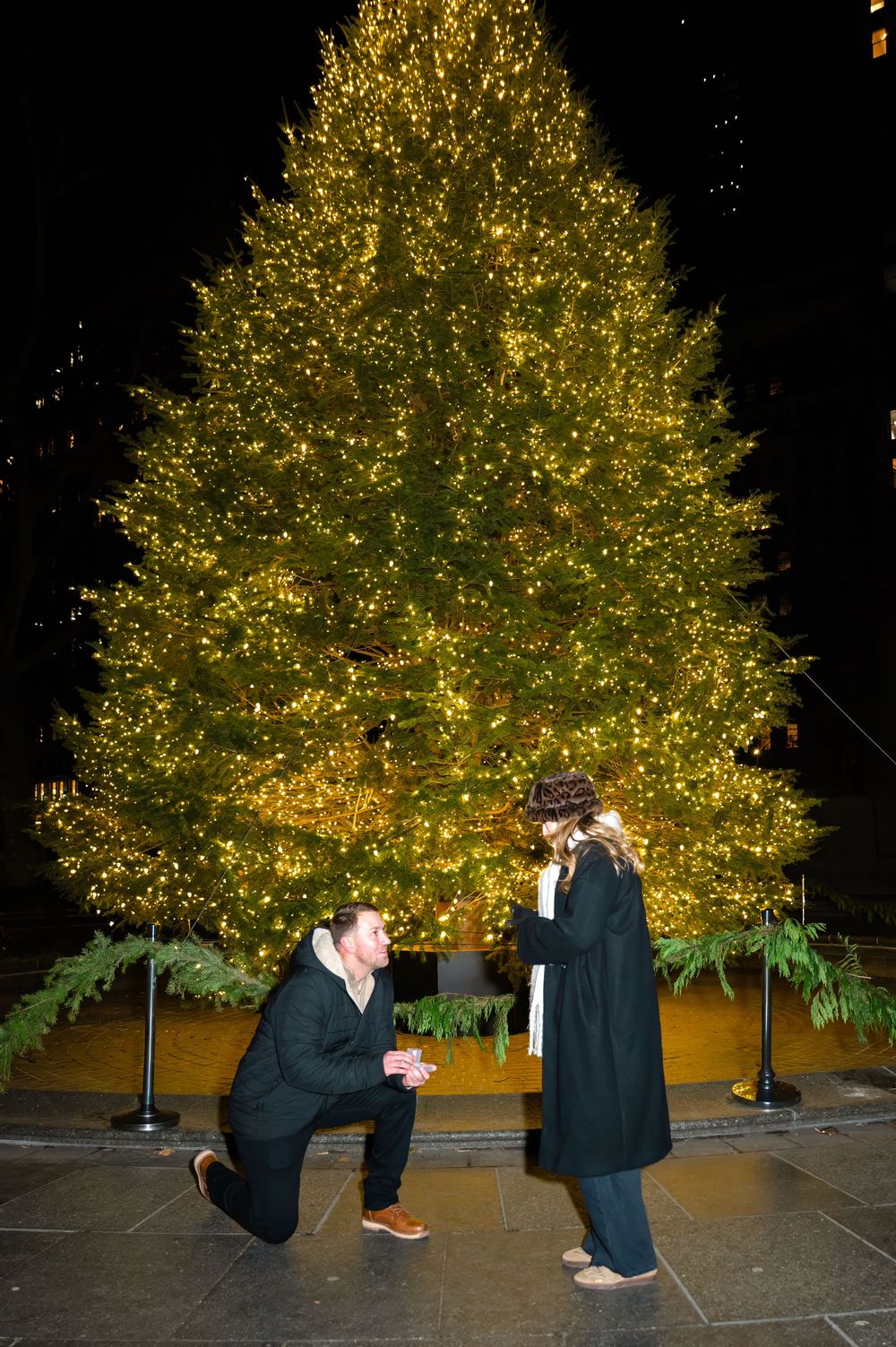 A romantic marriage proposal scene at night in front of a beautifully lit Christmas tree with golden lights in Madison Square Park in NYC.
