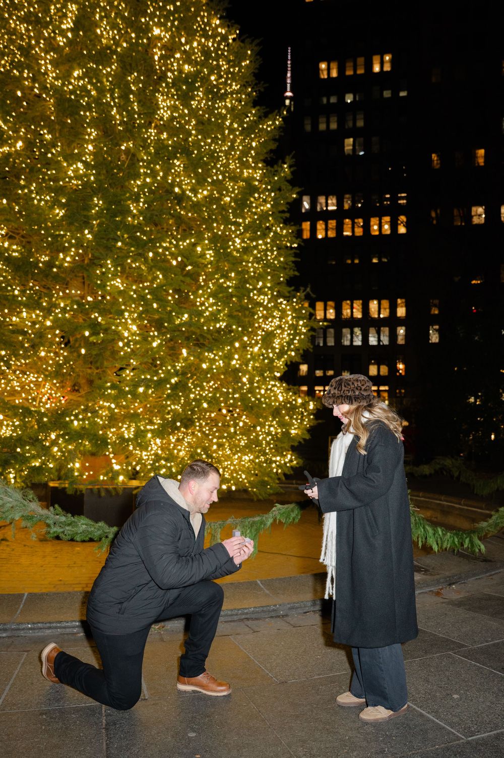 A couple sharing a special moment during a nighttime proposal in front of an illuminated tree with city buildings behind.