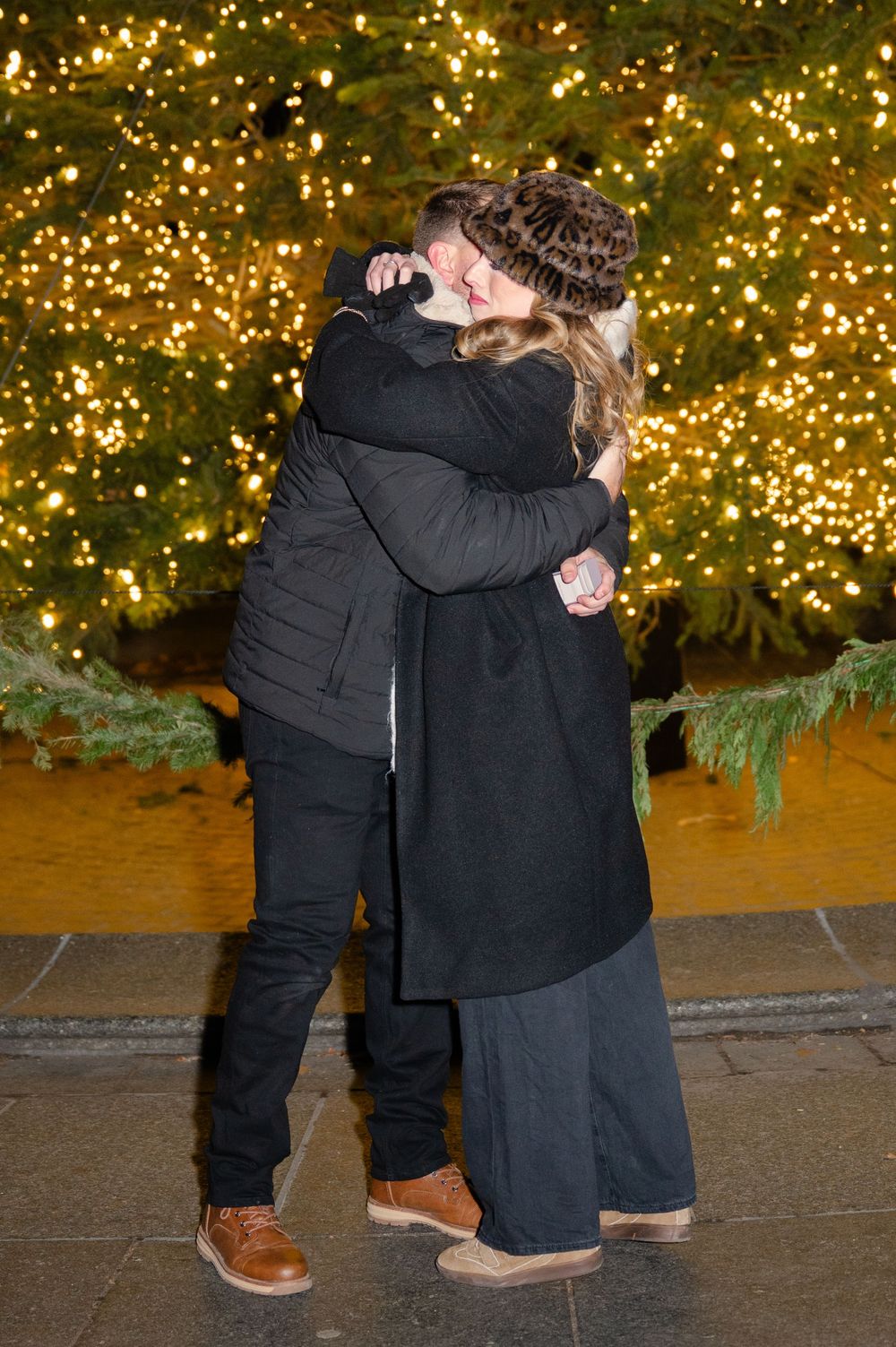 Two people in black winter coats hug while standing near a Christmas tree with twinkling golden lights in the background in NYC.