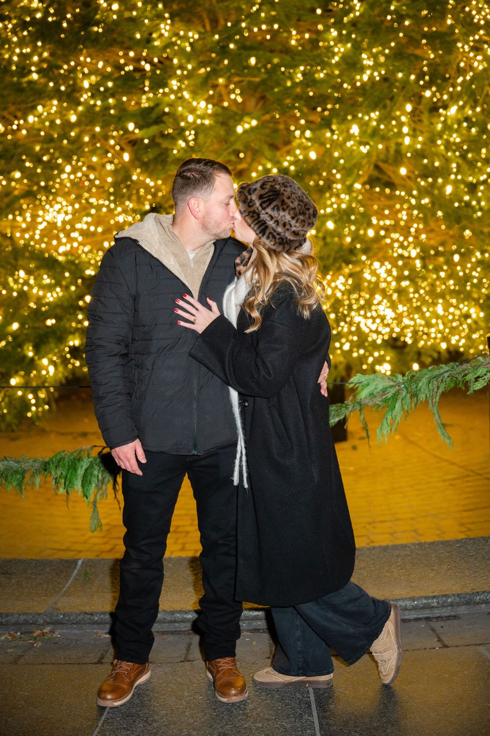 A couple sharing a romantic kiss against a backdrop of golden holiday lights in front of a tree during an NYC holiday proposal.