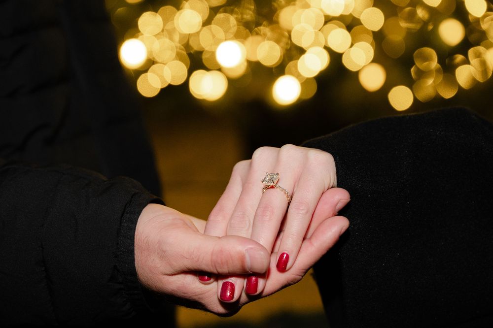 A hand with red nail polish holds a gold engagement ring against a blurred bokeh background of warm glowing lights.