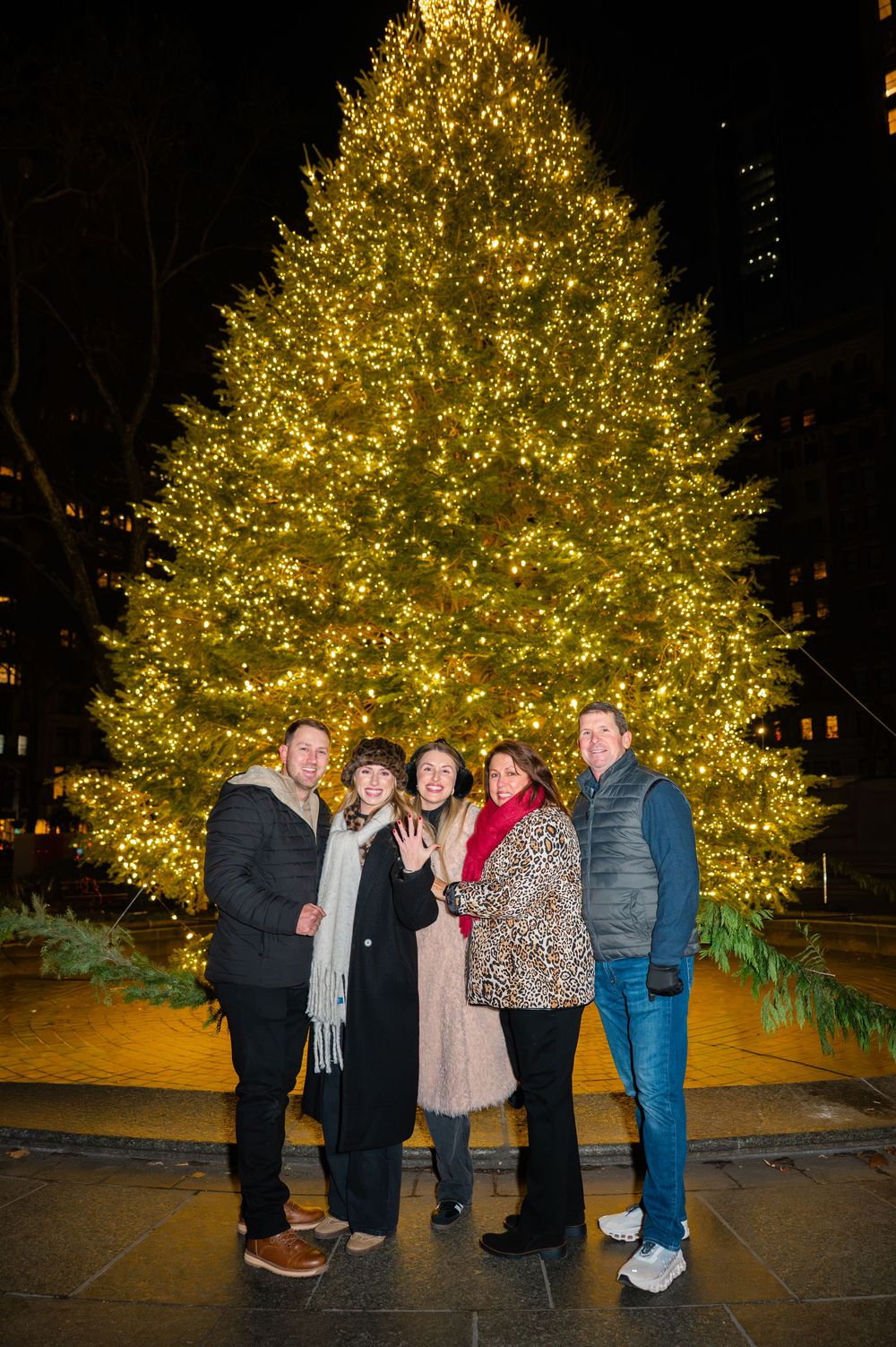 A group gathers in front of a beautifully lit Christmas tree glowing with warm golden lights at night.