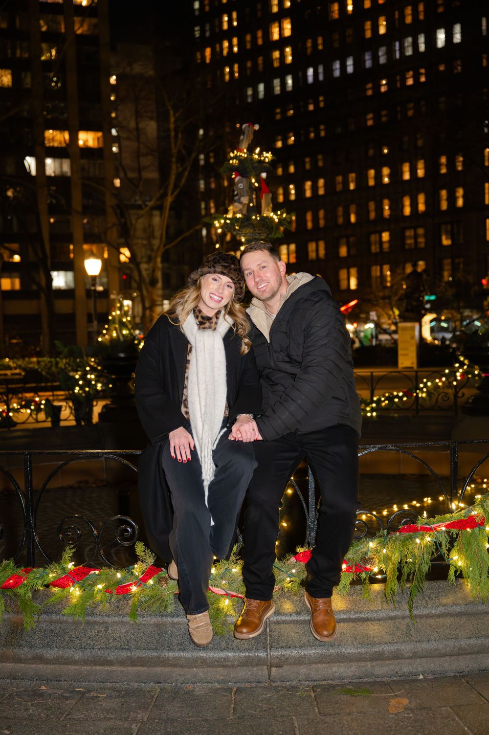 A couple laughs together while sitting on a decorated ledge with city buildings and holiday lights in the background.