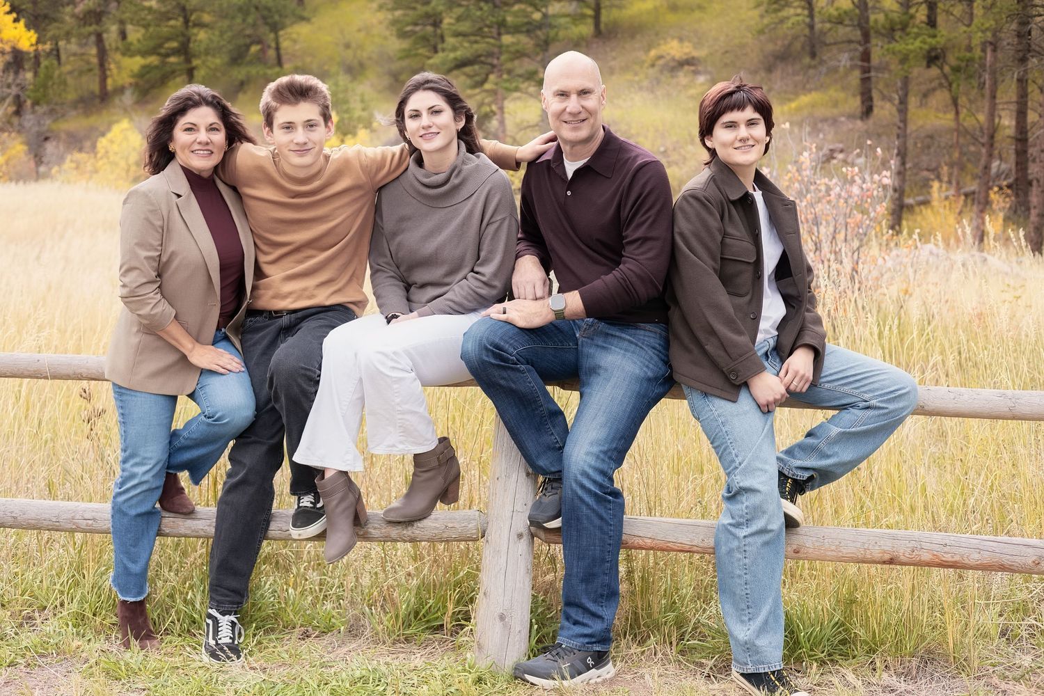 A group sits casually together outdoors on a wooden fence against an autumn forest backdrop.