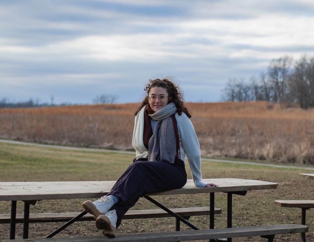 Cheerful young woman sitting on outdoor picnic table in open field on cloudy day.