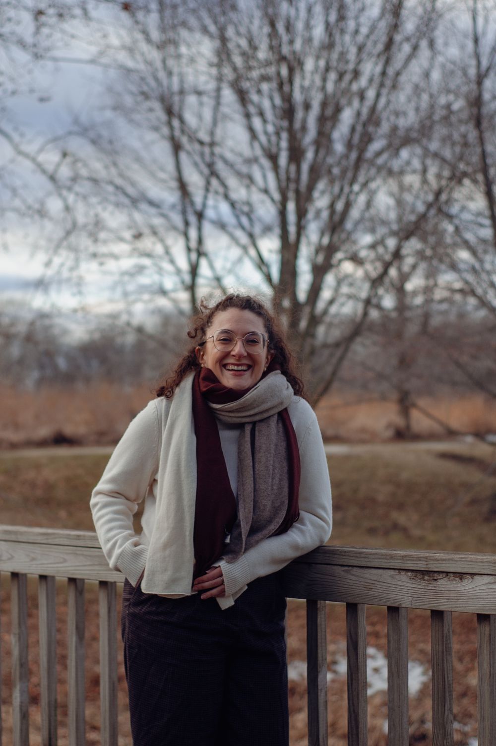 Smiling woman with curly hair wearing white jacket and layered scarves on wooden bridge.