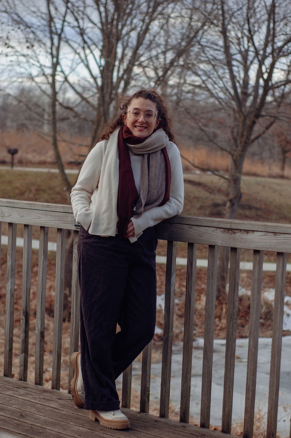 Full length portrait of smiling woman in white jacket and scarves leaning on wooden railing.