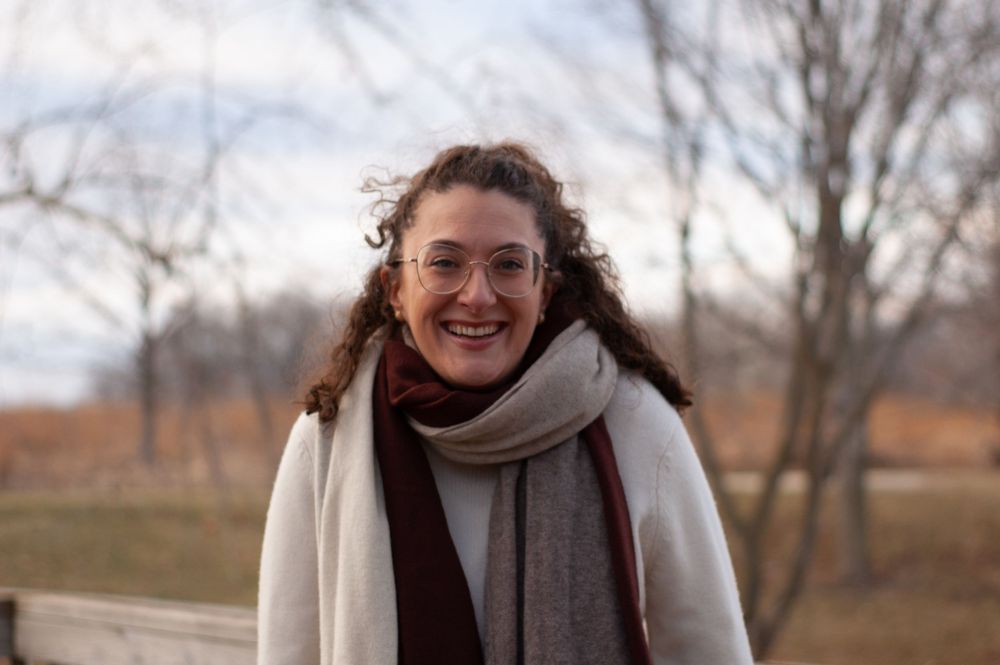 Close-up portrait of smiling curly-haired woman wearing glasses and layered scarves outdoors.