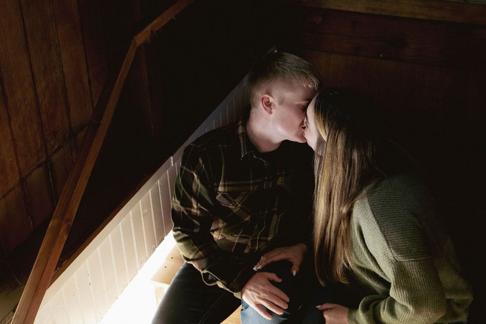 Romantic couple sharing a kiss in dimly lit wooden staircase of a barn.