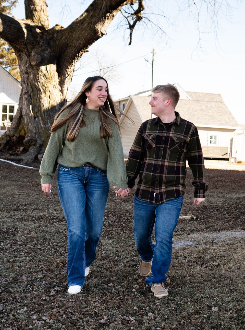 Happy young couple holding hands and walking outdoors near large bare tree in winter.