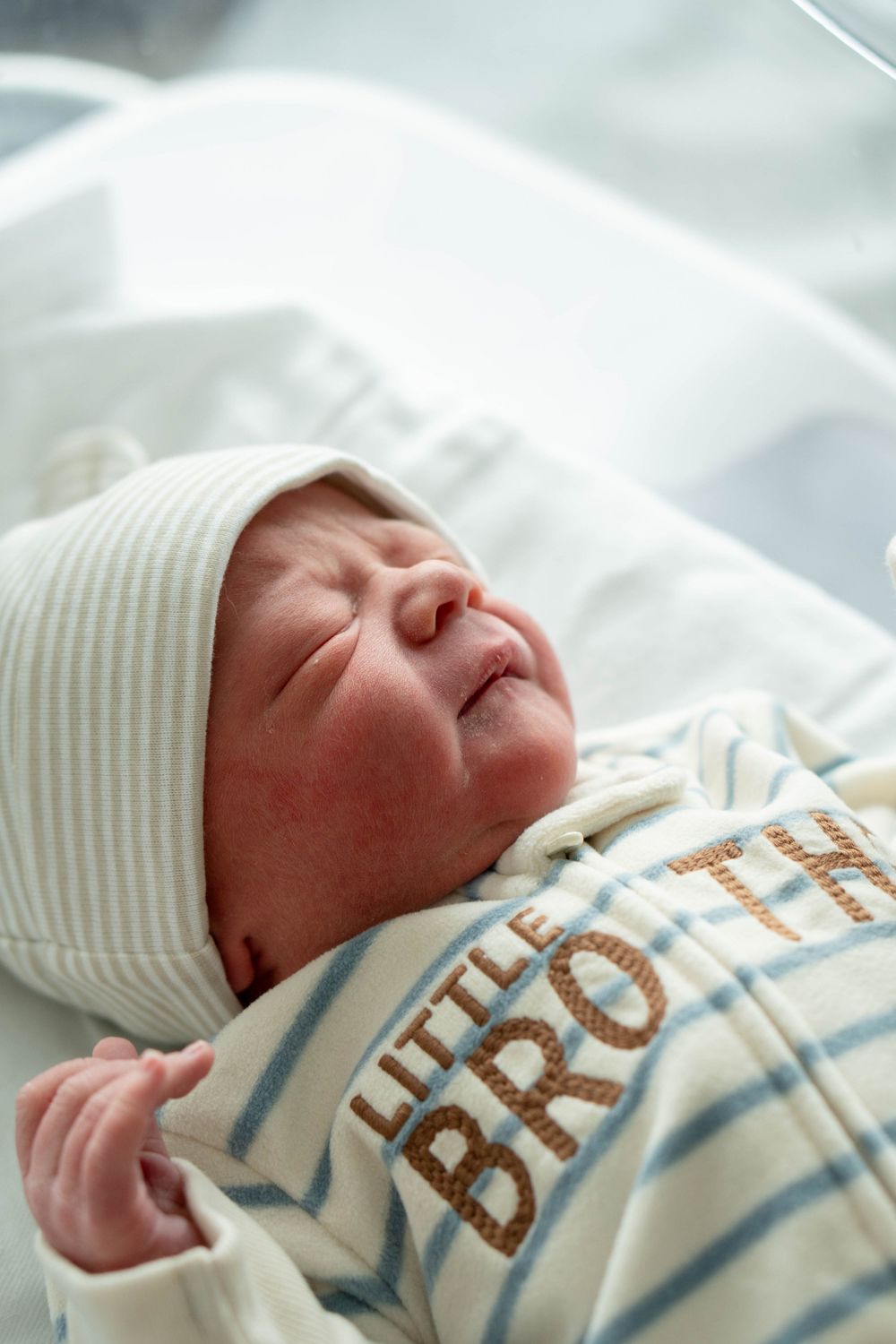 Newborn baby sleeping in hospital bassinet wearing a Little Brother striped onesie and hat.