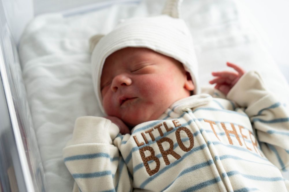 Peaceful newborn baby resting in hospital crib dressed in a Little Brother outfit and white hat.
