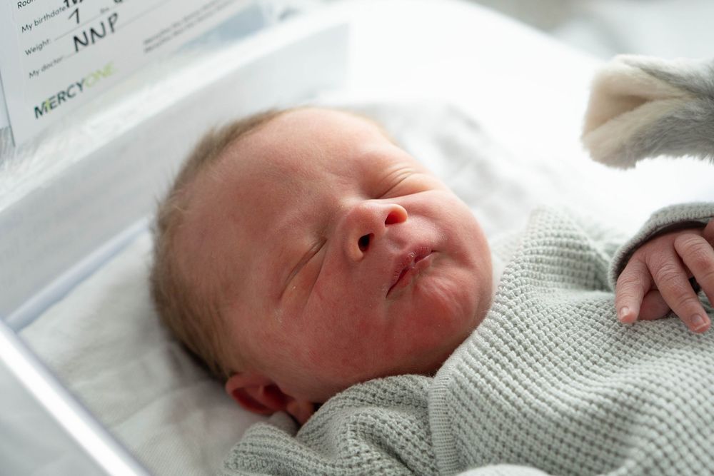 Newborn baby sleeping peacefully in hospital bassinet wrapped in soft green knit blanket.